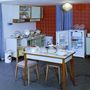 1962: fa-, üveg-, műanyag és lakkozott felületek gazdagítják a pompás összhatást, a kezdetleges konyhasziget a merészen a szoba közepét elfoglaló étkezőasztal is egyben